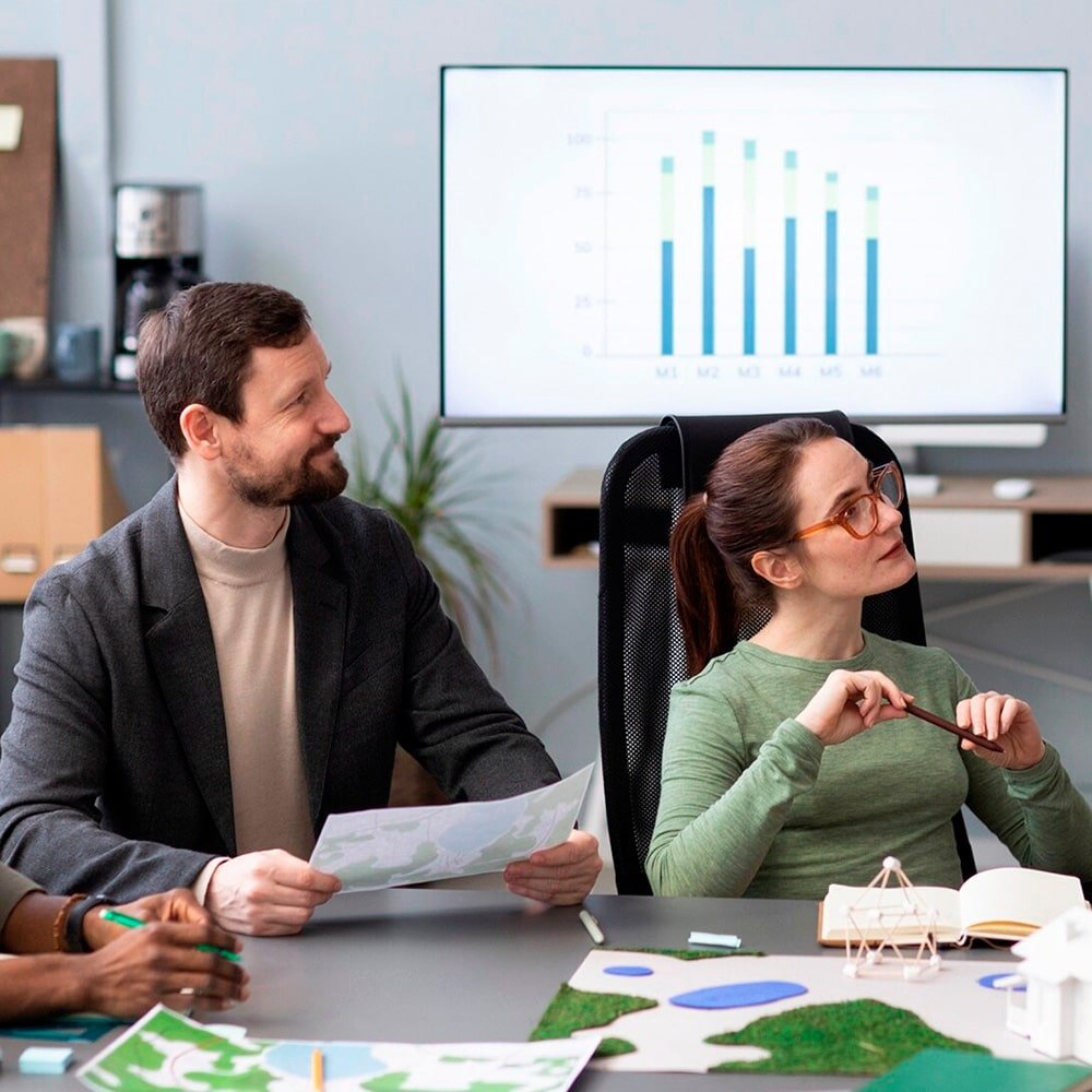 Dos personas en un ambiente laboral observan atentamente a alguien fuera de cuadro, con maquetas y anotaciones sobre la mesa.